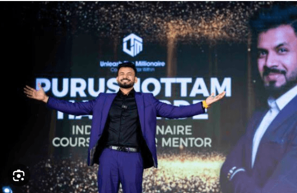 Smiling man in blue suit with arms outstretched on stage before a Purushottam branded screen.