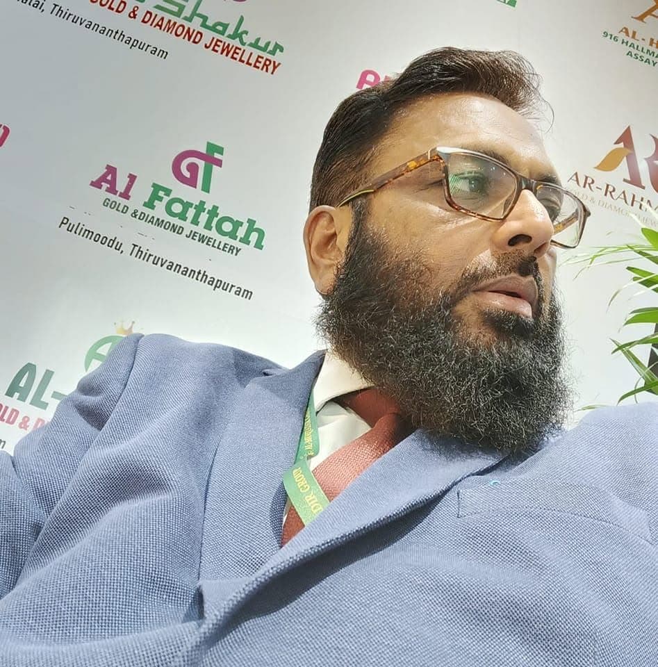 Bearded man in a blue blazer and glasses posing in front of jewelry brand logos.
